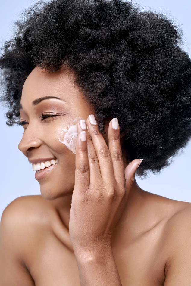 Woman with curly hair applying face cream, smiling with closed eyes. Her bare shoulders suggest skincare routine. Background is a soft blue.