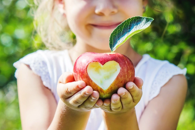 A smiling child holds a red apple with a heart-shaped bite. The apple has a green leaf on top. The scene exudes happiness and warmth.