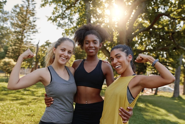 Three women in athletic wear smiling and flexing their arms in a park, with sunlight filtering through trees. The mood is joyful and empowering.